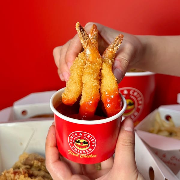 Hands dipping three crispy fried shrimp into a red cup of sauce from Cluck & Crispy Chicken.