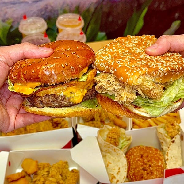 Hands holding a cheeseburger and a crispy chicken sandwich, with fries and wraps in boxes below.