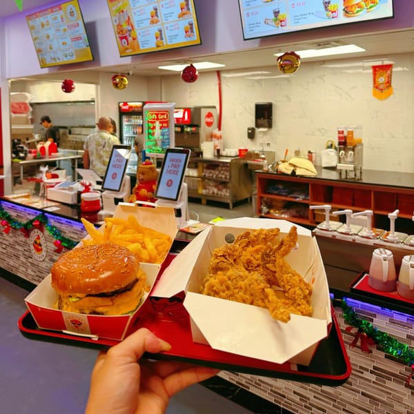 A hand holds a tray with a Cluck & Crispy chicken burger, fries, and fried chicken in a fast-food restaurant.