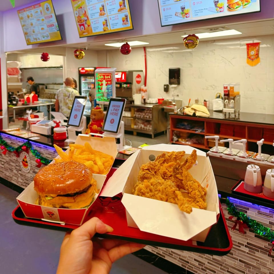 A hand holds a tray with a Cluck & Crispy chicken burger, fries, and fried chicken in a fast-food restaurant.