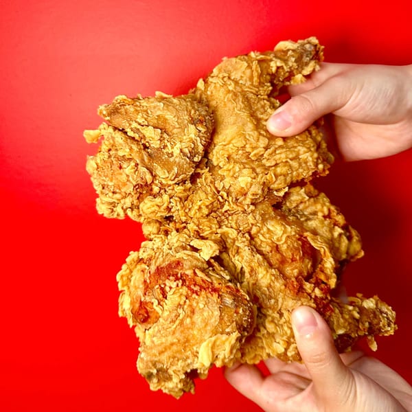 Two hands holding a large piece of golden-brown crispy fried chicken against a vibrant red background.