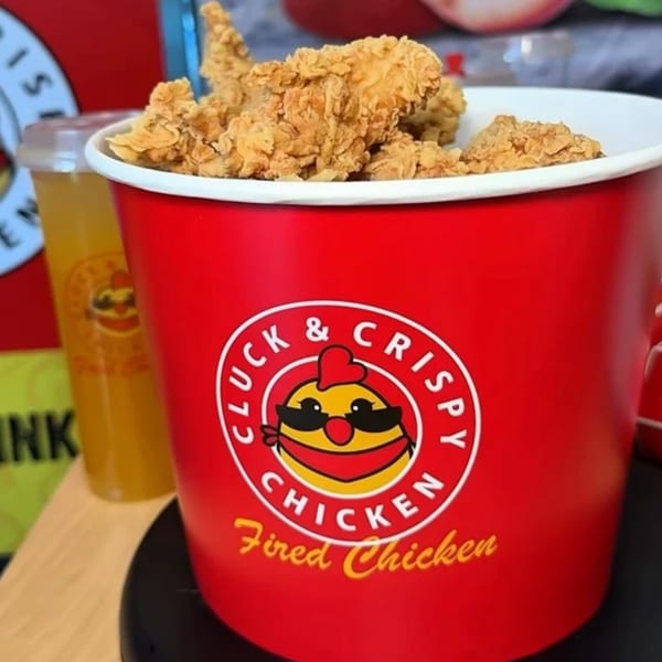 A red Cluck & Crispy Chicken bucket filled with golden fried chicken, next to a drink.
