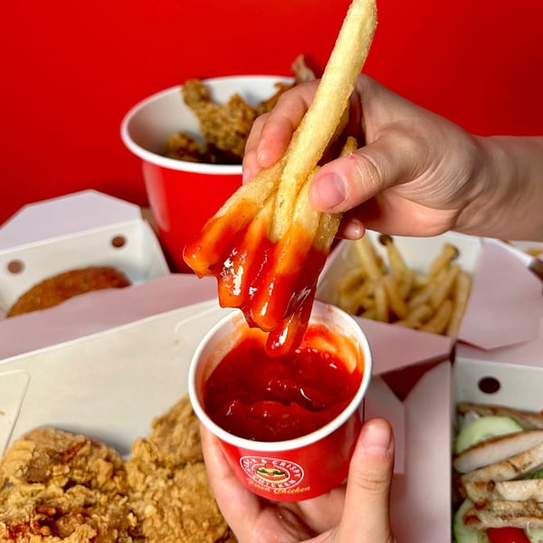 Hand dipping Cluck & Crispy Chicken fries into a red cup of ketchup, surrounded by fast-food items.