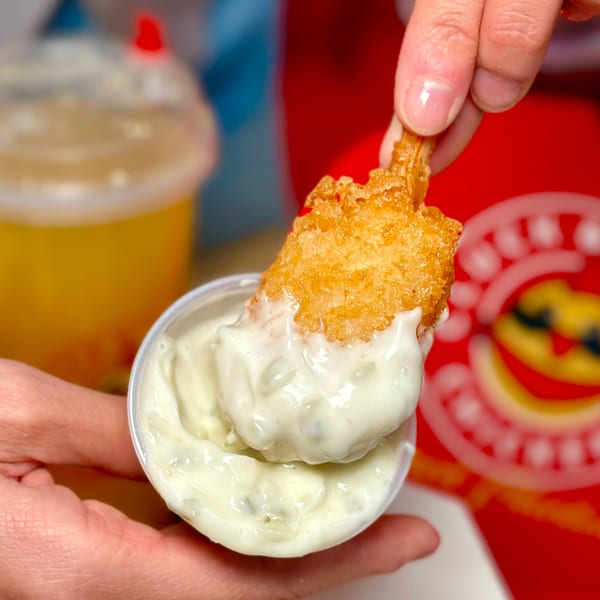 A hand dips a crispy Cluck & Crispy chicken tender into a small cup of creamy white sauce.