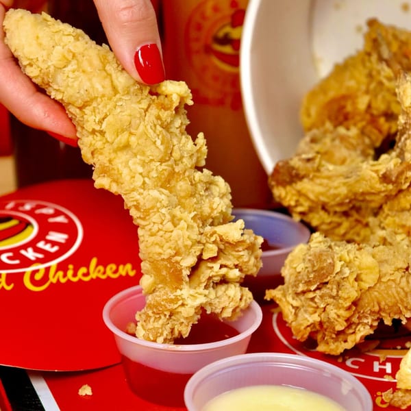 A hand dips a crispy Cluck & Crispy chicken tender into a small cup of red sauce.