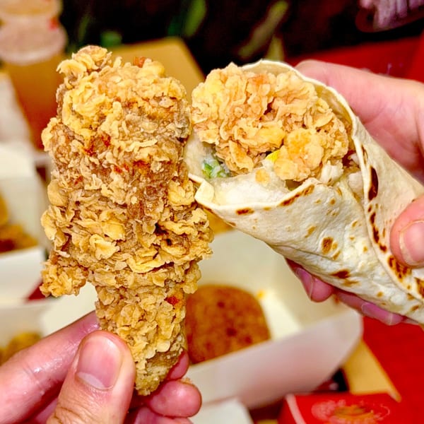 Hands holding a crispy fried chicken piece and a chicken wrap from Cluck & Crispy Chicken.